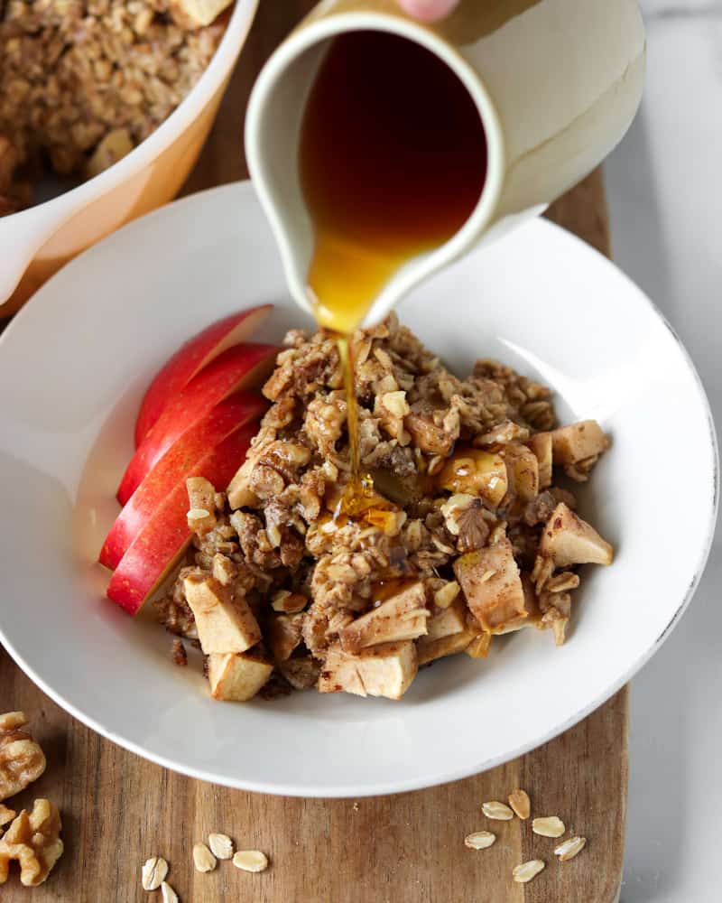 A bowl of warm apple cinnamon baked oatmeal, with a pitcher of maple syrup being poured on top