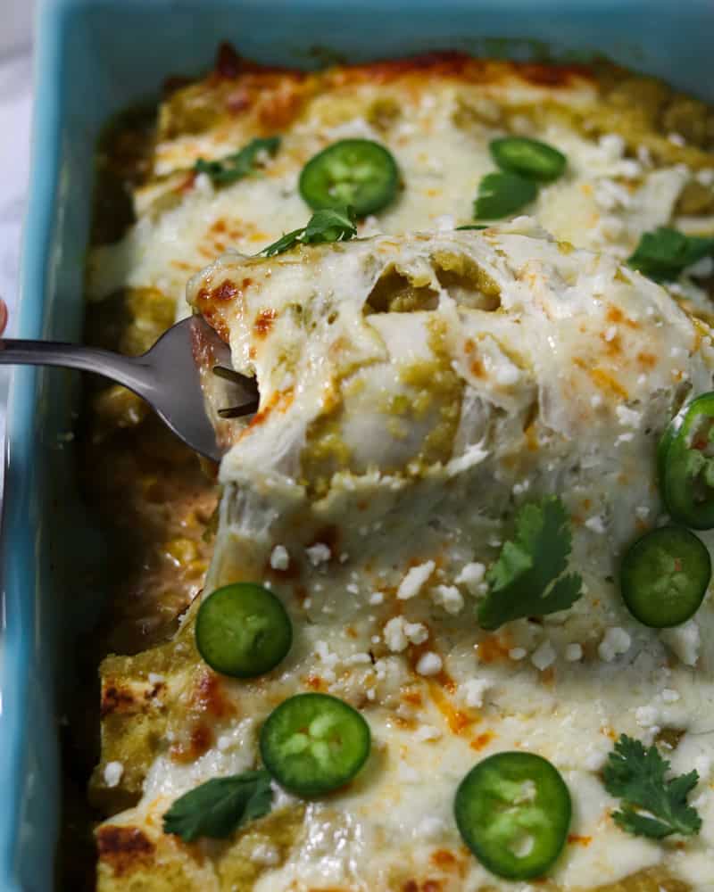 A spatula pulling street corn enchiladas out of the pan, stretching the melty cheese on top