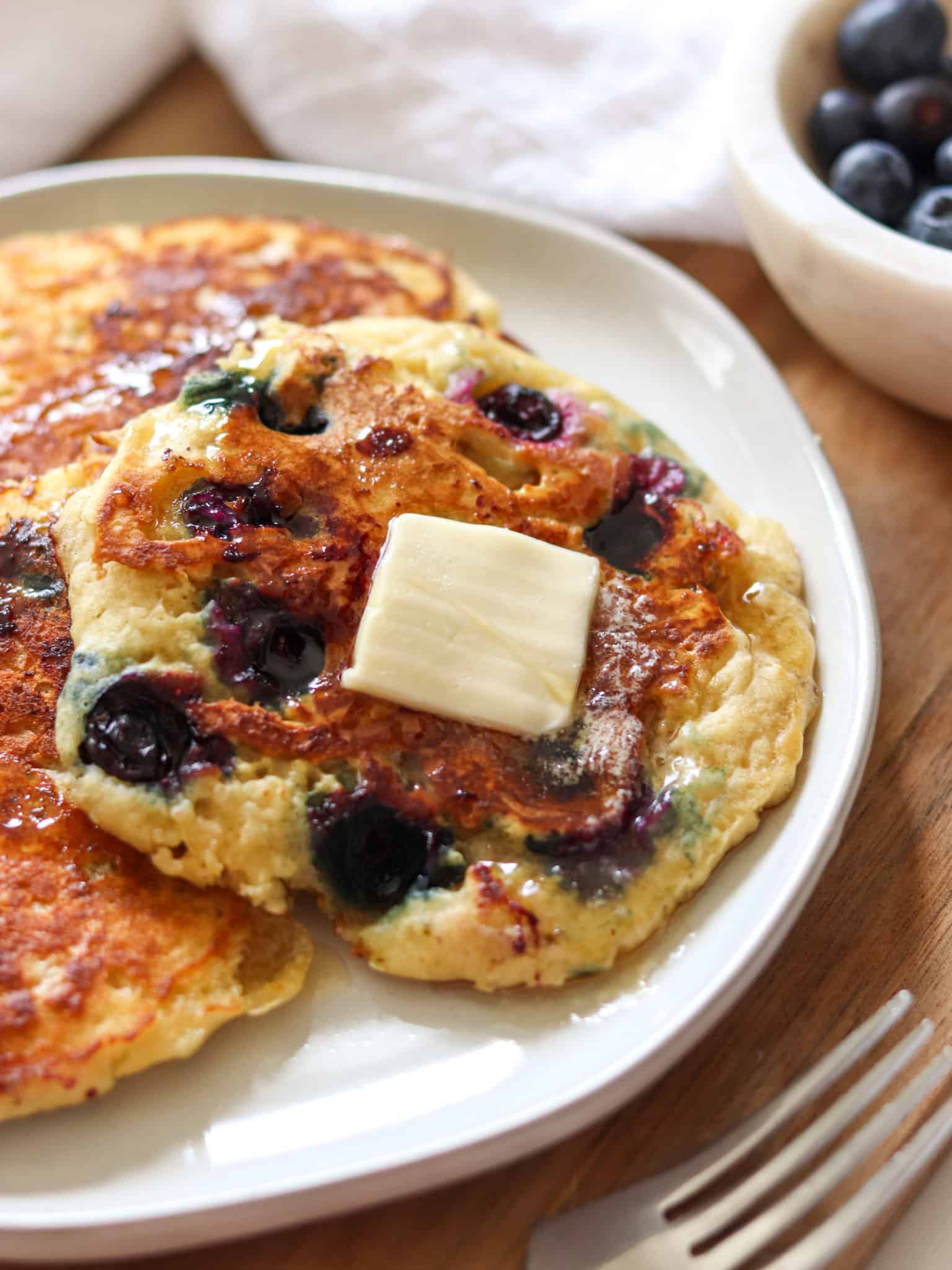 Blueberry pancakes on a plate with a pat of butter and maple syrup on top, and a bowl of fresh blueberries in the background