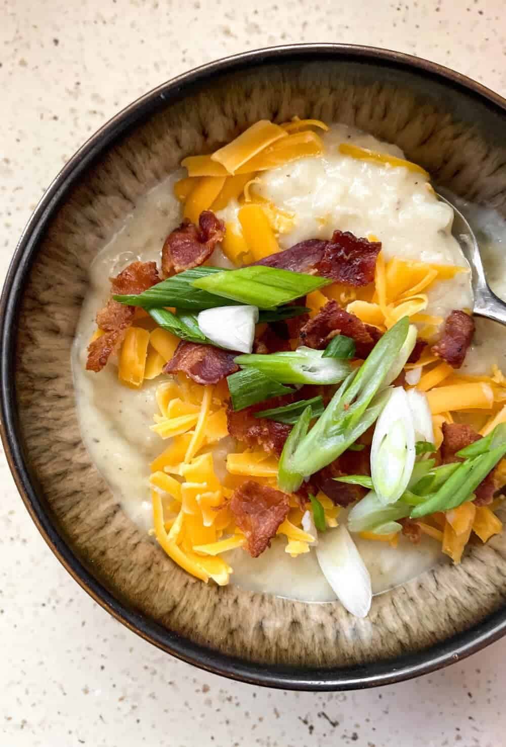 A bowl full of creamy loaded potato soup topped with melty cheddar cheese, crisp bacon and sliced green onions, with a spoon scooping out some of the soup