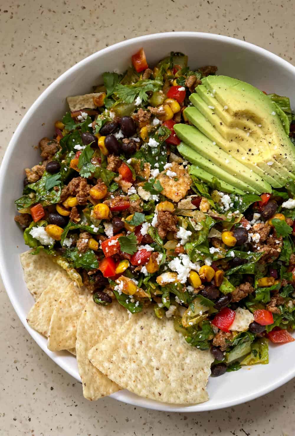 A bowl heaping with fresh, colorful chopped taco salad (lettuce, peppers, beans, corn, turkey, cheese, and more), topped with sliced avocado and tortilla chips