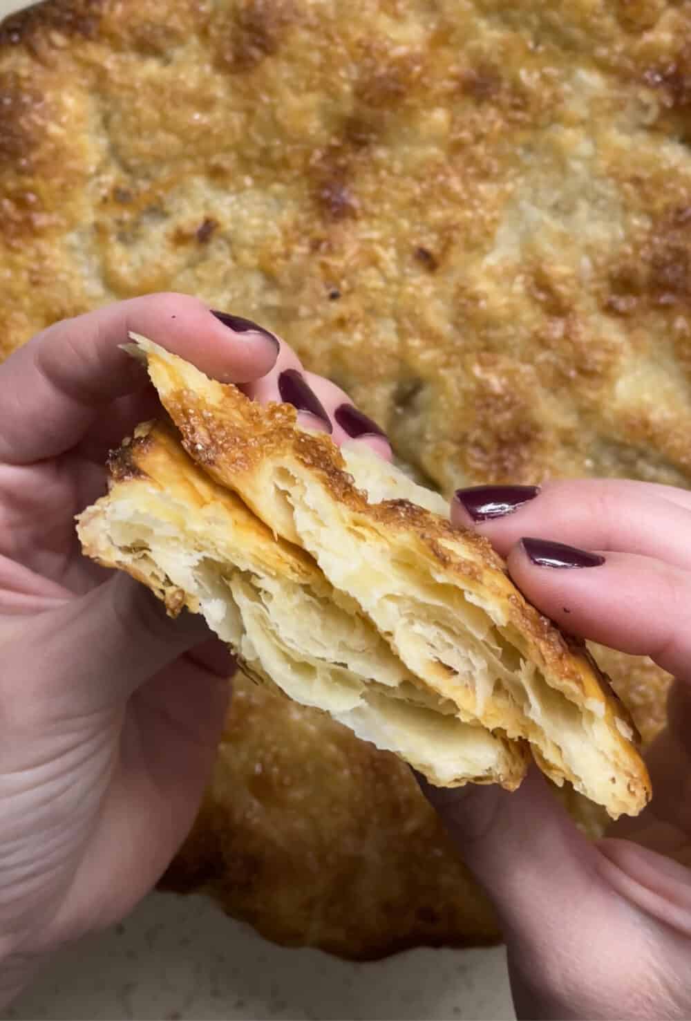 Hands holding a piece of pie crust, torn in half to show the buttery, flaky layers inside