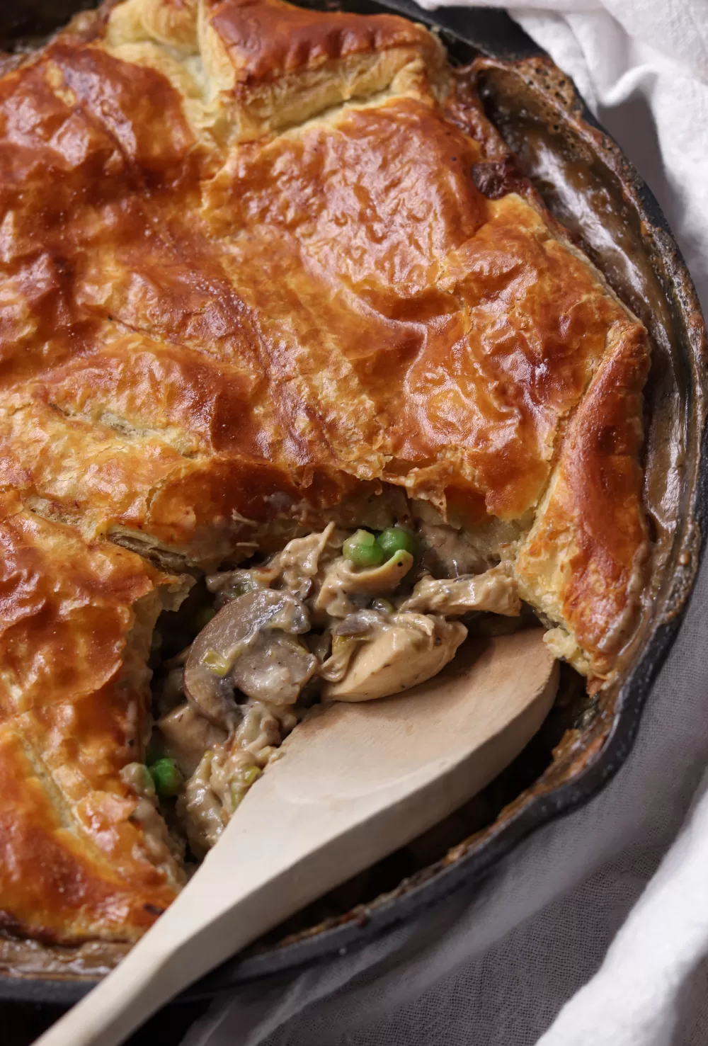 A puff pastry chicken pot pie with a wooden spoon in it, having taken out a scoop to reveal the chicken and mushroom filling inside.