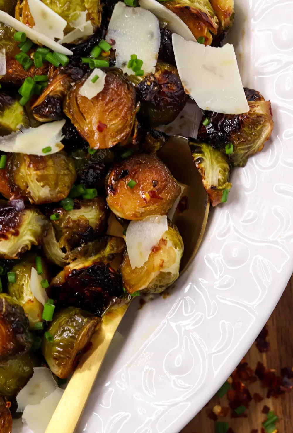 A close-up of hot honey Brussels sprouts, topped with shaved Parmesan cheese and chopped chives, on a serving platter with a gold spoon.