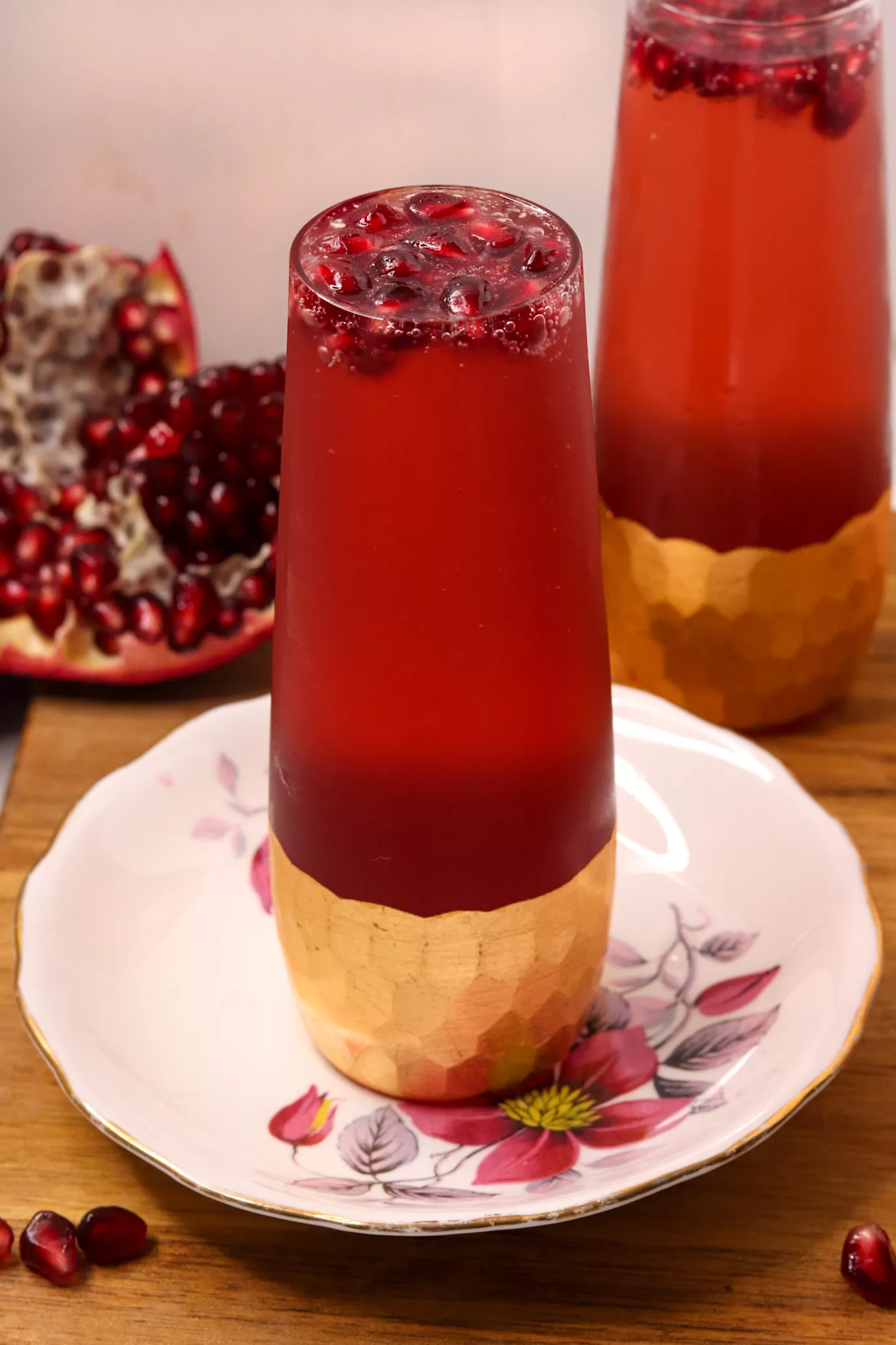 Two pomegranate French 75s in gilded champagne flutes on a wooden board. One of them is on an antique china plate.